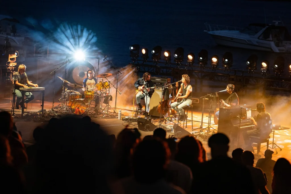 A rock band performing on a dramatically lit outdoor stage at night, with spotlights, fog, and a crowd in the foreground, representing the energy and pacing a writer aims to bring to their manuscript