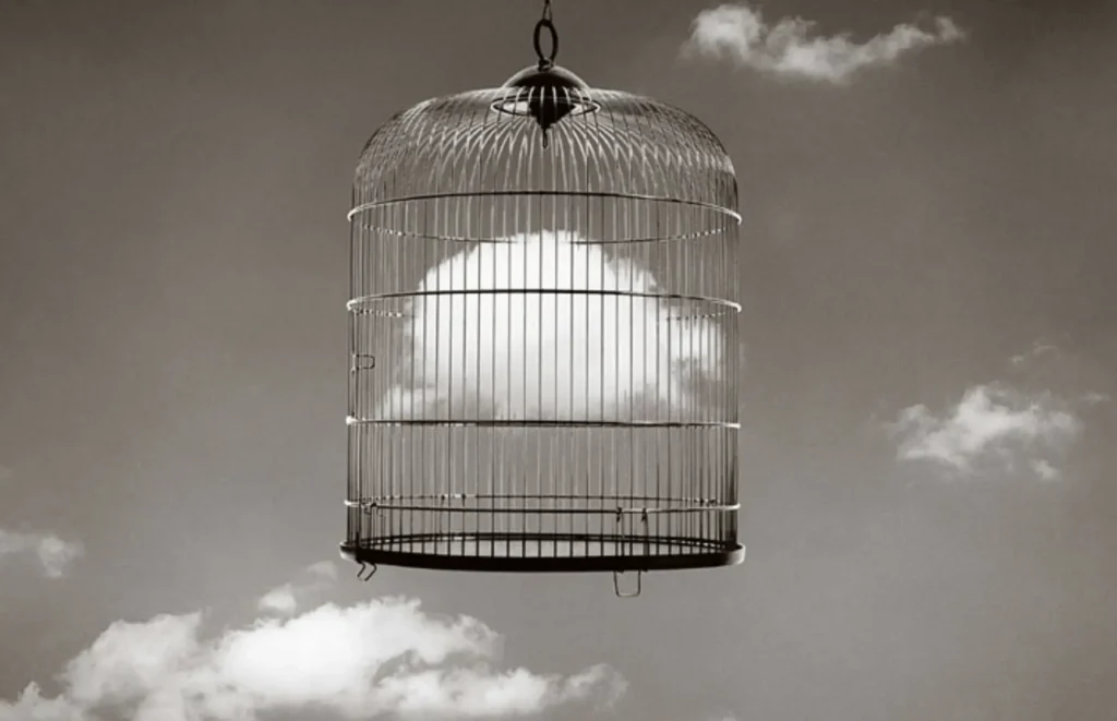 A surreal black-and-white photograph of an empty birdcage suspended in a cloudy sky, symbolizing the feeling of creative confinement and the longing for freedom that writers experience during periods of brain fog and life disruption
