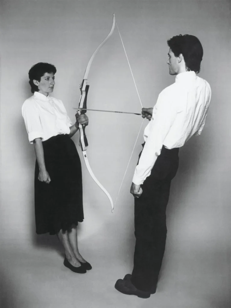 A black-and-white photograph of a man and woman facing each other, Marina Abramovic and Ulay, jointly holding a taut bow and arrow between them, representing the tension between a writer's cherished idea and the courage required to finally commit it to the page