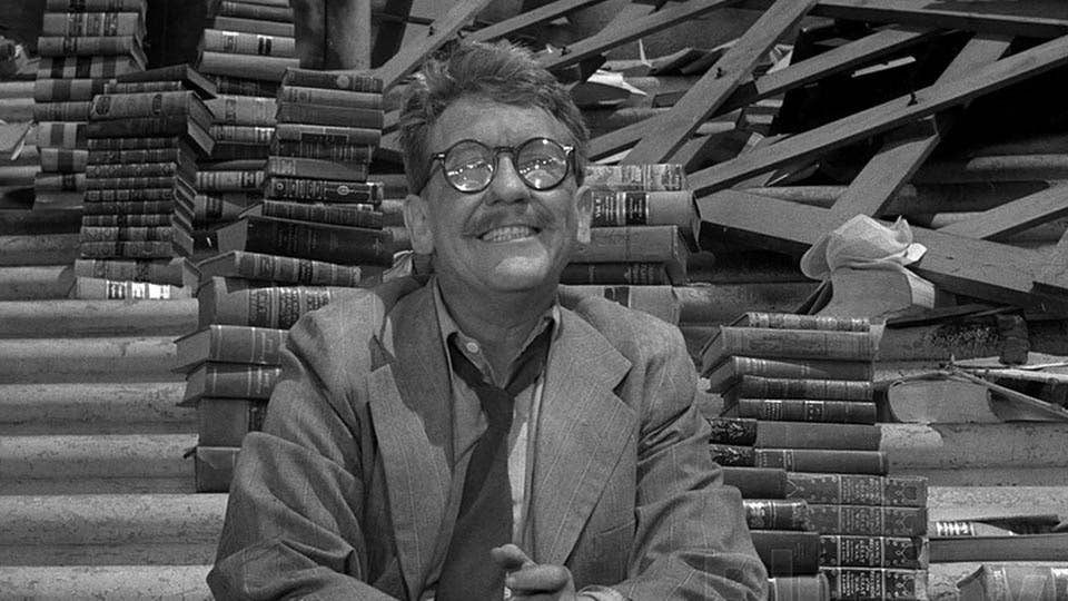 A black-and-white still from the Twilight Zone episode "Time Enough at Last," showing a bespectacled man surrounded by towering stacks of books in a post-apocalyptic setting, representing the writer's fantasy of finally having unlimited time to read and write — and the reality that never quite arrives