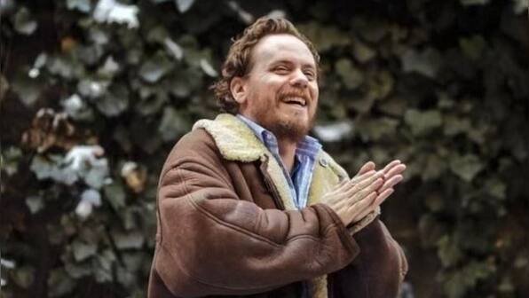 A portrait of late indie publisher and editor Giancarlo DiTrapano laughing and clapping outdoors against a backdrop of dark ivy, wearing a brown shearling jacket. As an iconic editorial force who urged writers to embrace their rawest instincts, his legacy embodies the Hewes House approach to manuscript development: helping authors stop searching for an external voice and bravely commit to the authentic one they already possess.